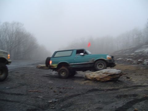 1993 Ford Bronco - A teal 1993 Ford Bronco navigates a rocky trail on a foggy day, with another vehicle in the background.