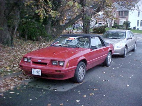 1991 Ford LTD Crown Victoria - A red convertible Mustang is parked on a street with fallen leaves, alongside another car.