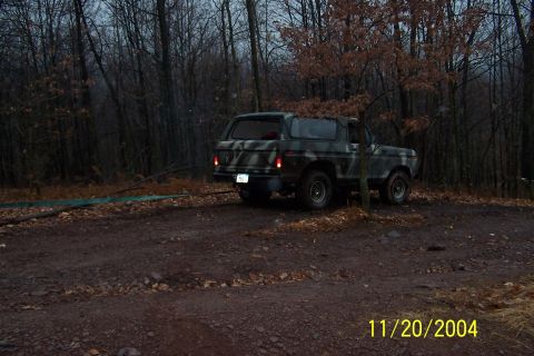 1979 Ford Bronco - A camouflaged Ford Bronco is parked on a dirt road surrounded by trees.