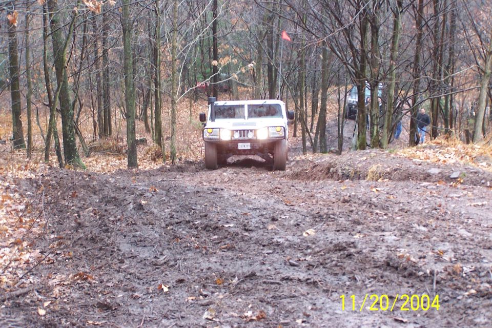White SUV with snorkel and off-road tires navigates a muddy trail in a wooded area. Another vehicle is visible behind it