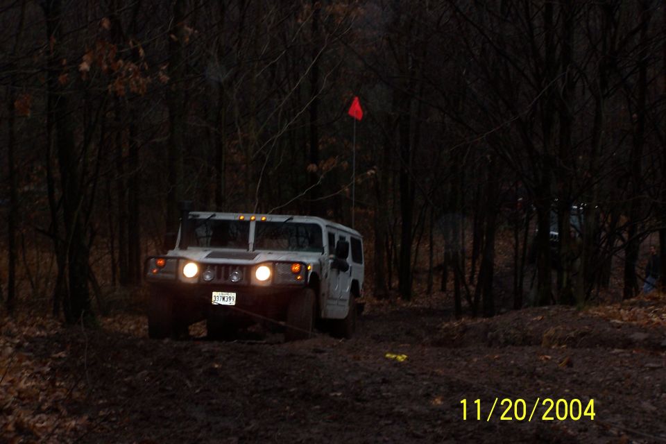 White SUV driving on a muddy trail in a dark forest, with headlights on and a red flag visible