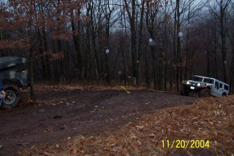 1979 Ford Bronco - Two off-road vehicles traverse a challenging muddy terrain in a wooded area during overcast weather.