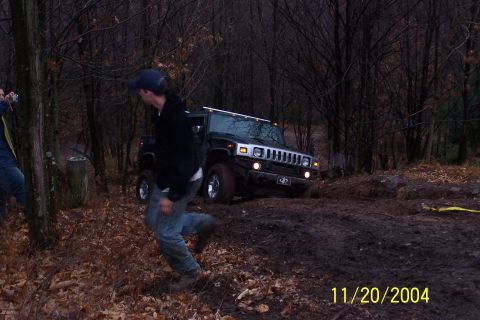 1979 Ford Bronco - A black Hummer H2 navigates a muddy trail through a wooded area, with a person running in the foreground.