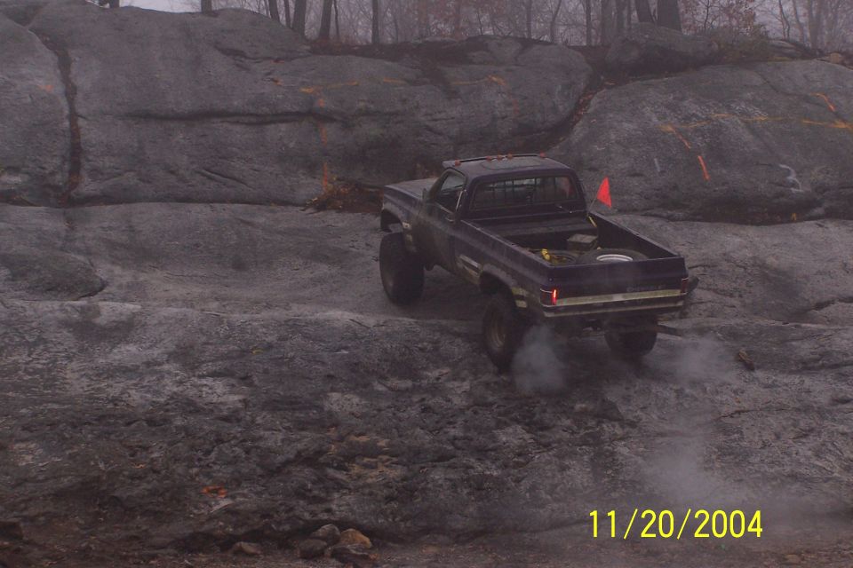 Dark blue pickup truck with lifted suspension driving over rocks, emitting smoke from rear tire