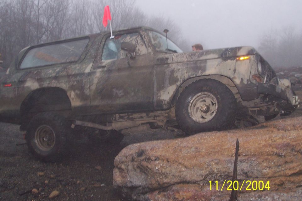 Mud-covered 1978 Ford Bronco on rocks, red flag on roof