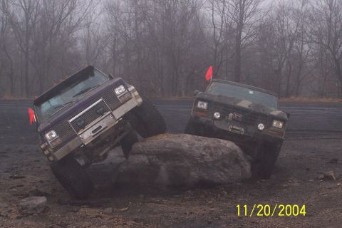 1979 Ford Bronco - Two trucks are shown navigating over a large rock during an off-road excursion on a foggy day.
