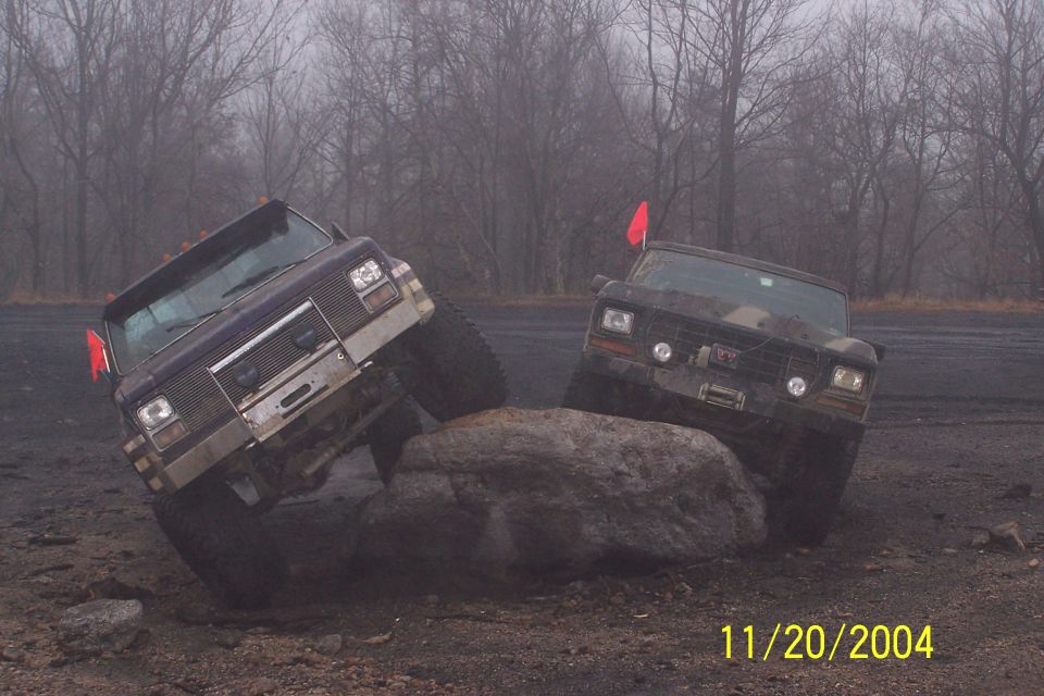 Two trucks are off-roading, one purple and one dark, navigating over a large rock. The purple truck has a chrome grille and bumper