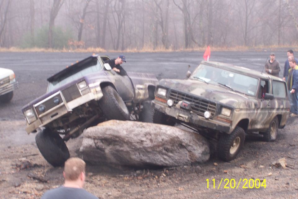 Two modified SUVs are attempting to drive over a large rock. One is purple and white, the other is camouflaged. A person is visible in the driver's seat of the purple vehicle