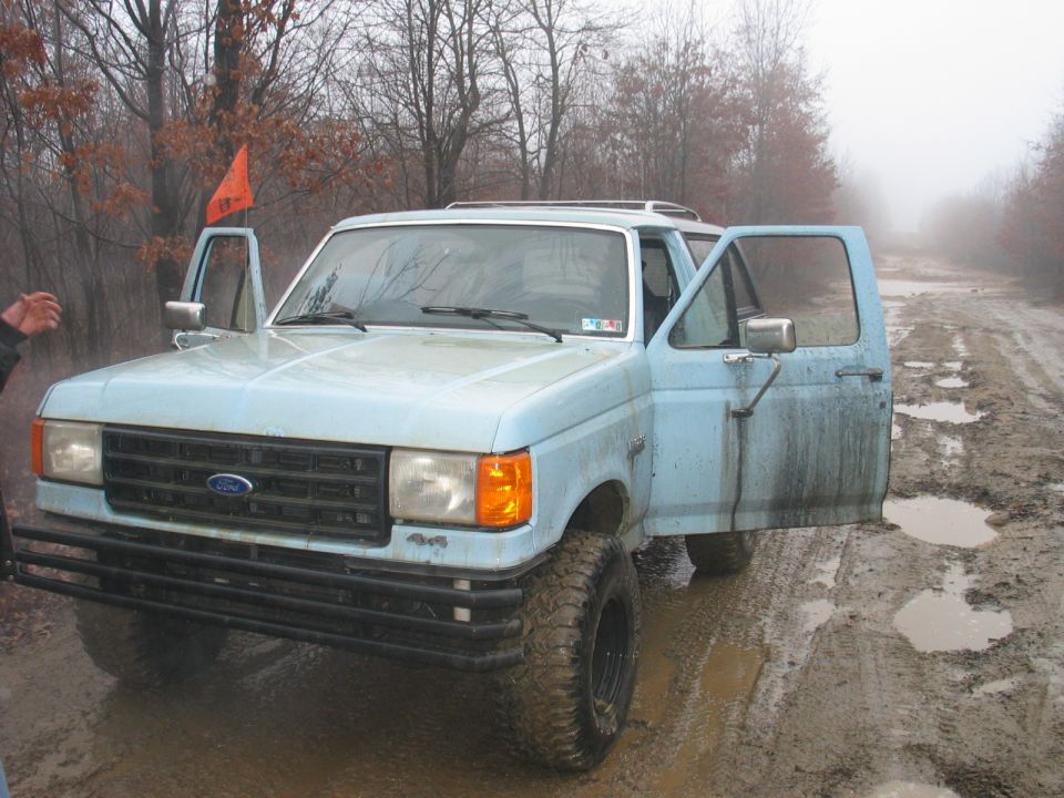 Light blue pickup truck, doors open, parked on a muddy, wooded road. Orange flag on driver's side
