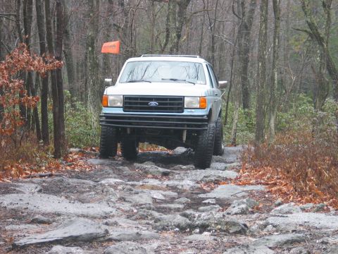 1995 Ford Bronco - A light blue Ford Bronco navigates a challenging rocky path through a wooded area.