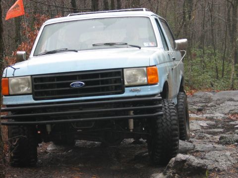 1995 Ford Bronco - This light blue Ford Bronco, likely from the late 1980s, navigates a challenging rocky trail with its prominent bull bar and large tires.