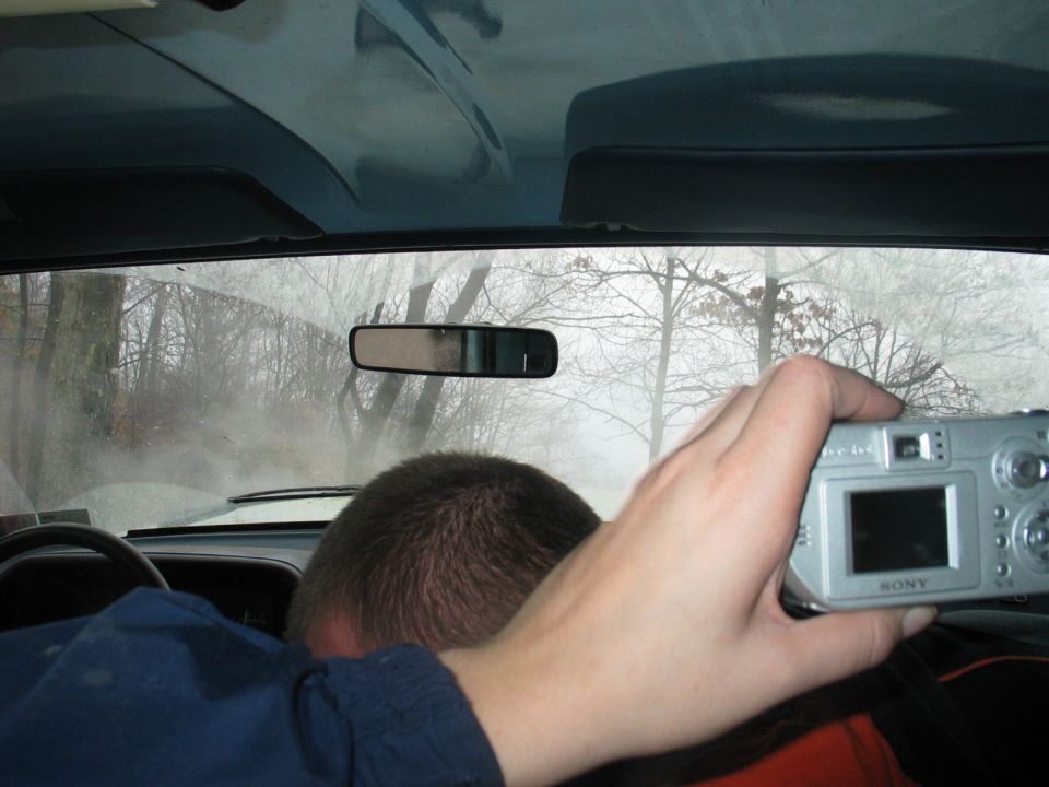 Interior view from a vehicle, looking through the windshield at a foggy, wooded area. A person's head is visible in the foreground, and another person's hand holds a silver Sony digital camera, taking a picture out the window