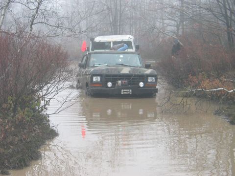 1995 Ford Bronco - Two trucks navigate a flooded, wooded area on an overcast day.