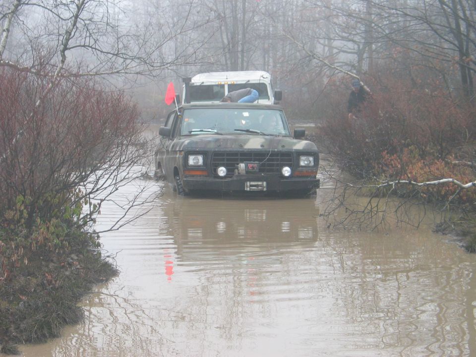 Camouflaged Ford F-Series truck driving through muddy water, with a white truck behind it