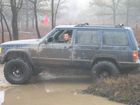 1995 Ford Bronco - A blue Jeep Cherokee XJ navigates a muddy off-road trail, its tires splashing through a large puddle.