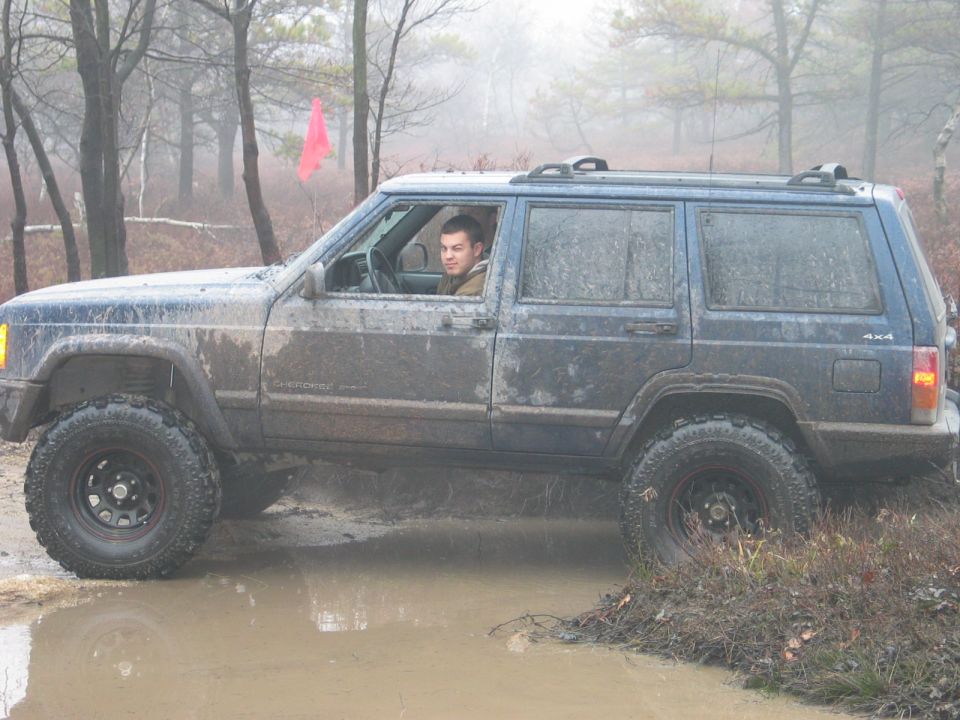 Blue Jeep Cherokee XJ side view, covered in mud, driving through a muddy puddle