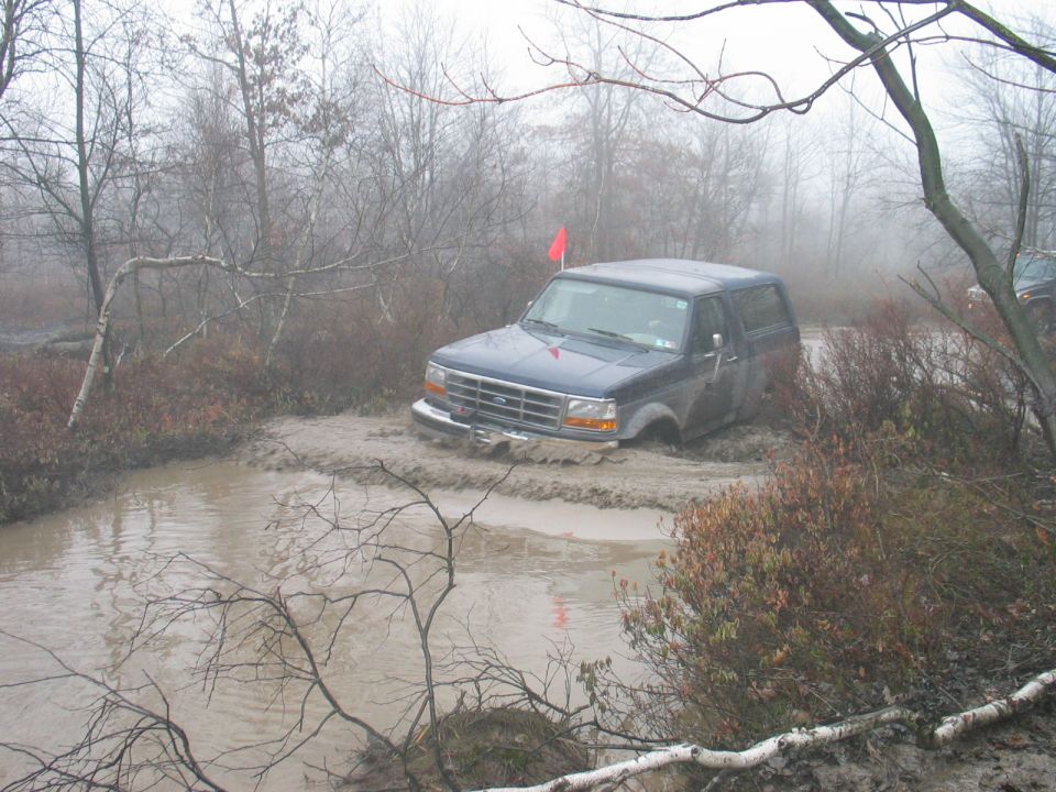 Dark blue 1995 Ford Bronco driving through a muddy trail with trees and fog in the background