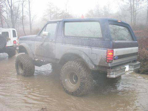 1995 Ford Bronco - A lifted Ford Bronco navigates through muddy water in a wooded area.