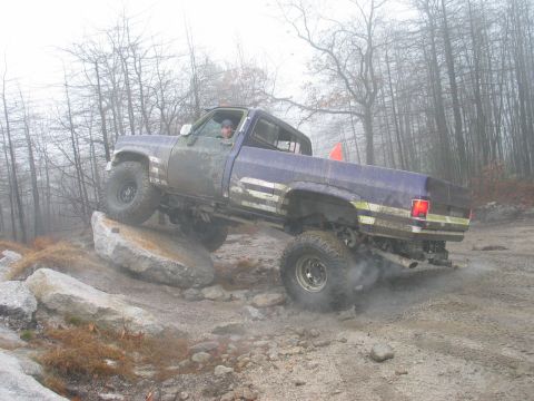 1995 Ford Bronco - A purple pickup truck navigates a challenging off-road course, with one wheel on a large rock and dust kicking up from the tires.