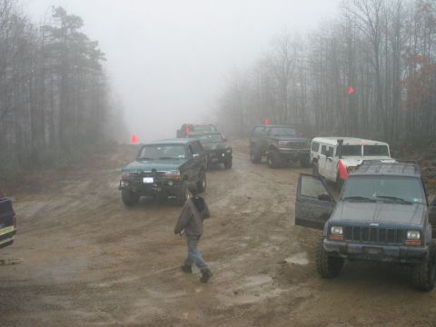 1995 Ford Bronco - Multiple off-road vehicles are gathered on a muddy path surrounded by trees on a foggy day.