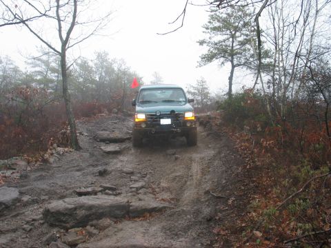 1995 Ford Bronco - A green SUV drives through a challenging, muddy, and rocky terrain on an overcast day.