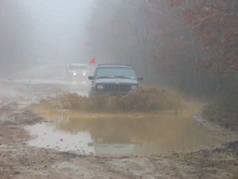 1995 Ford Bronco - Two off-road vehicles navigate a muddy trail on a foggy day, kicking up water and dirt.