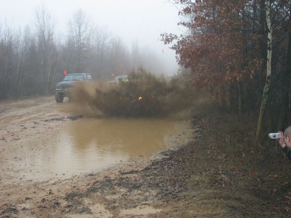 Dark-colored Ford Bronco driving through a muddy puddle, kicking up a large splash. Another vehicle is visible behind it
