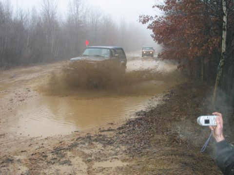 1995 Ford Bronco - Two SUVs navigate a muddy off-road trail, kicking up large plumes of water and dirt.