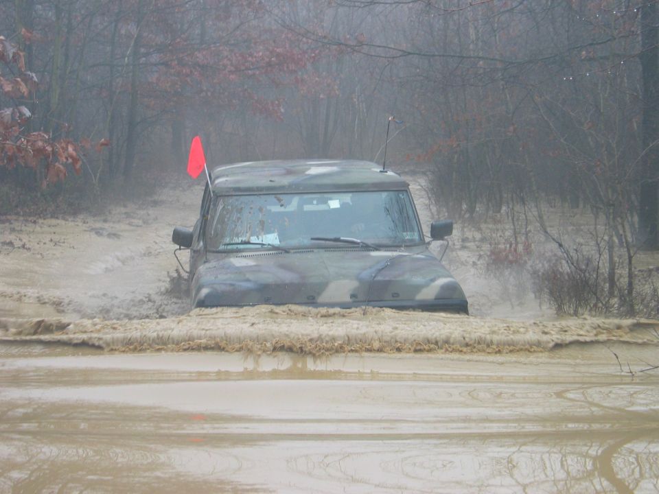 Camouflaged military SUV driving through deep mud and water, red flag on antenna