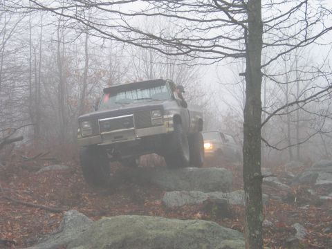 1995 Ford Bronco - Two off-road trucks traverse a rocky, fog-shrouded path in a forest.
