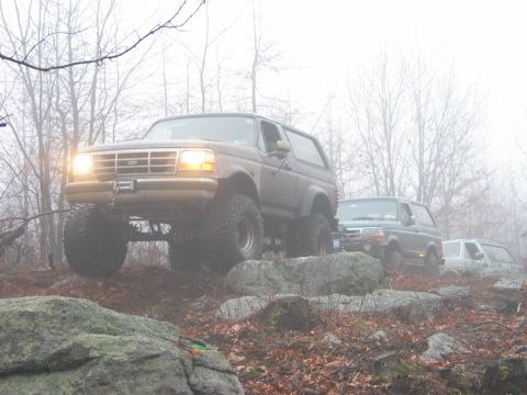 1995 Ford Bronco - Multiple Ford Broncos navigate a rocky, wooded trail on a foggy day.