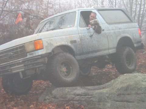 1995 Ford Bronco - A light blue Ford Bronco is shown off-roading over large rocks, with a driver visible inside.