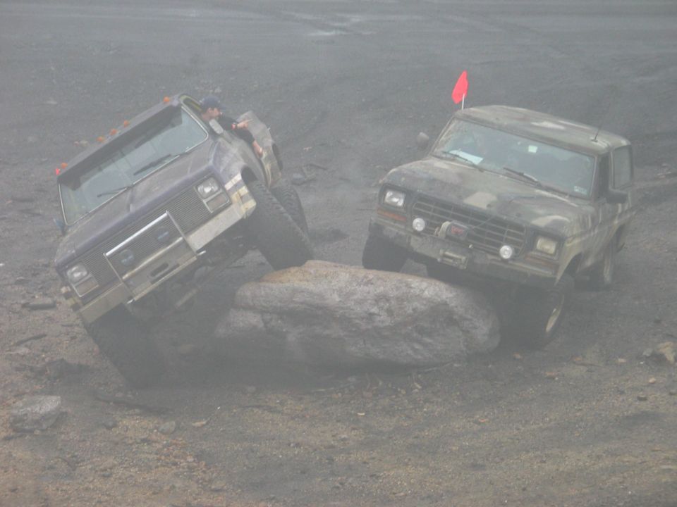Two older model Ford Broncos, one purple and one camouflage, climbing over a large rock on a dirt trail