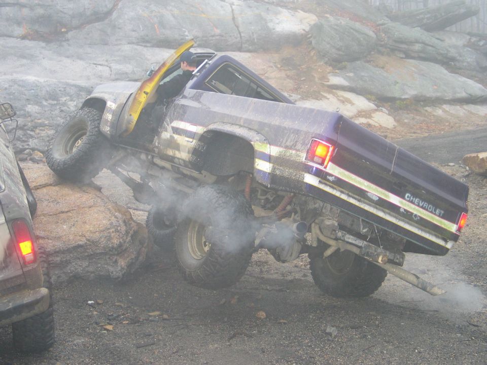 Pickup truck climbing over rocks with smoke coming from exhaust. Another vehicle is to the left