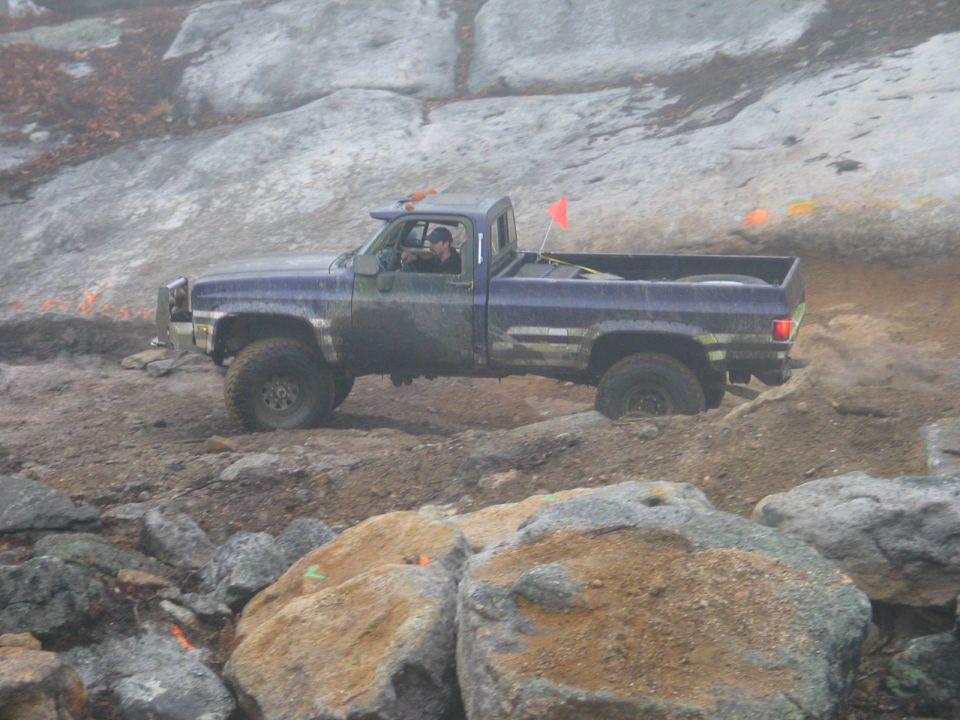 Purple pickup truck driving over rocks and dirt. Driver wears a cap
