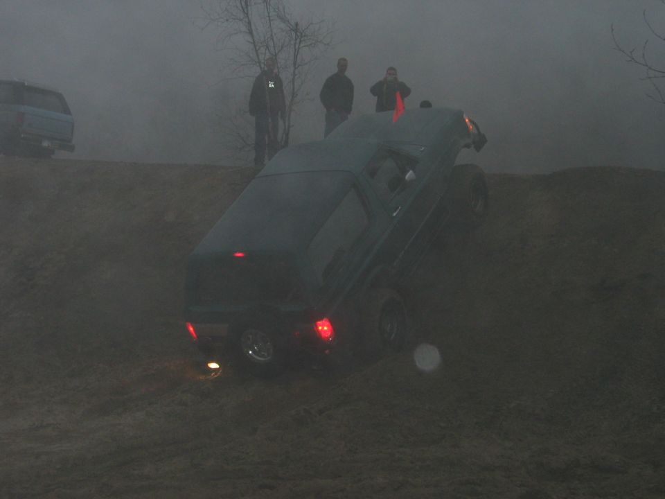 Dark SUV climbing a muddy hill in fog with people watching. Another SUV is visible in the background