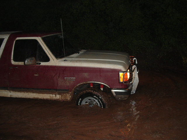 Maroon and white Ford Bronco driving through muddy water at night