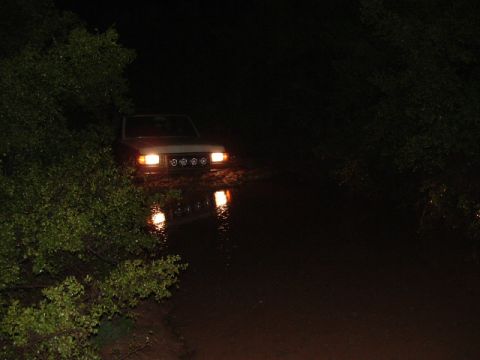 1990 Ford Bronco - A 1990 Ford Bronco navigates through murky water at night, its headlights illuminating the path ahead.