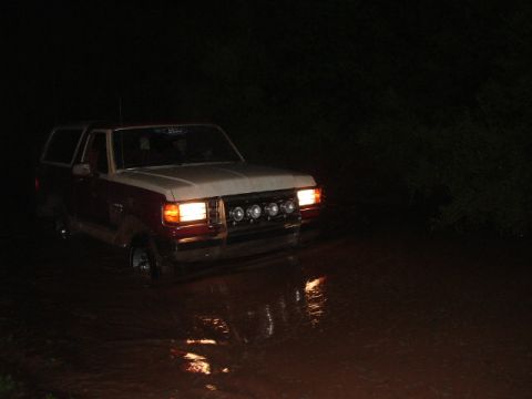 1990 Ford Bronco - A two-tone Ford Bronco navigates through murky water in the dark, its headlights illuminating the path ahead.