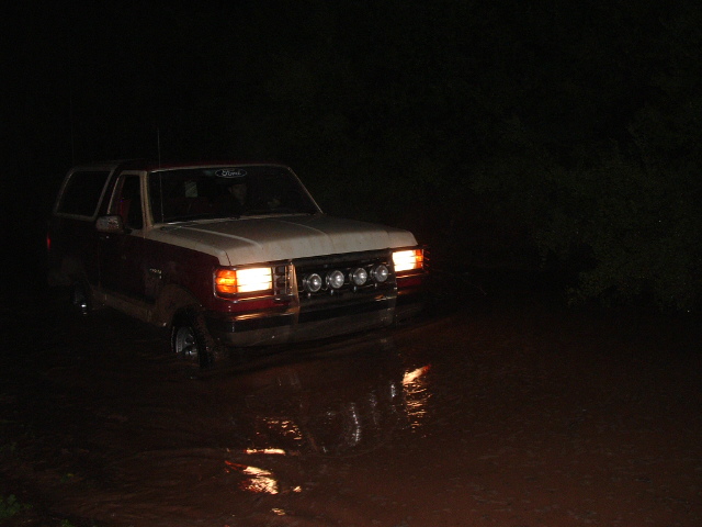 Two-tone Ford Bronco driving through water at night, front grille with auxiliary lights