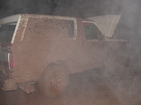 1990 Ford Bronco - Passenger-side profile of a full-size two-door SUV buried under a uniform coat of dried brown dust, with the hood raised and dust still han…