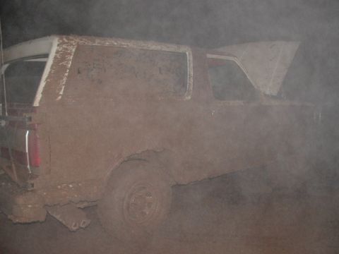 1990 Ford Bronco - Side profile of a full-size two-door SUV completely coated in dried brown dust and mud, photographed in a thick cloud of hanging dust. The…