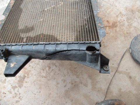 1990 Ford Bronco - A removed radiator lying on a stained concrete garage floor, photographed along the black plastic side tank where a long crack runs through…