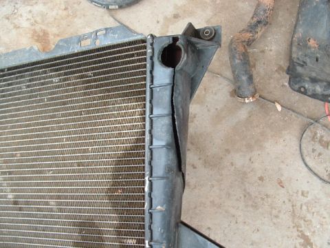 1990 Ford Bronco - A removed radiator lying face-up on a concrete garage floor, showing the aluminum core fins and the black plastic side tank with its upper…