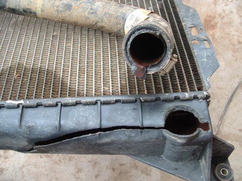 1990 Ford Bronco - Close-up of a removed radiator lying on concrete with its black plastic end tank split wide open along the crimp seam next to the lower hos…
