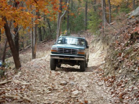 1995 Ford Bronco - A green SUV navigates a rough, off-road trail surrounded by autumn foliage.