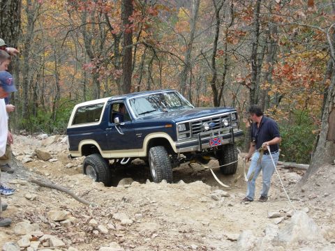 1995 Ford Bronco - A blue and white Ford Bronco is seen off-roading on a rocky, wooded trail.