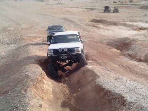 1985 Ford Bronco - A white SUV, likely from the 1990s, tackles a challenging off-road course with other vehicles in the background.