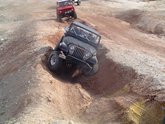 Black soft-top Jeep crawling down a deep dirt ravine off-road, red Jeep behind