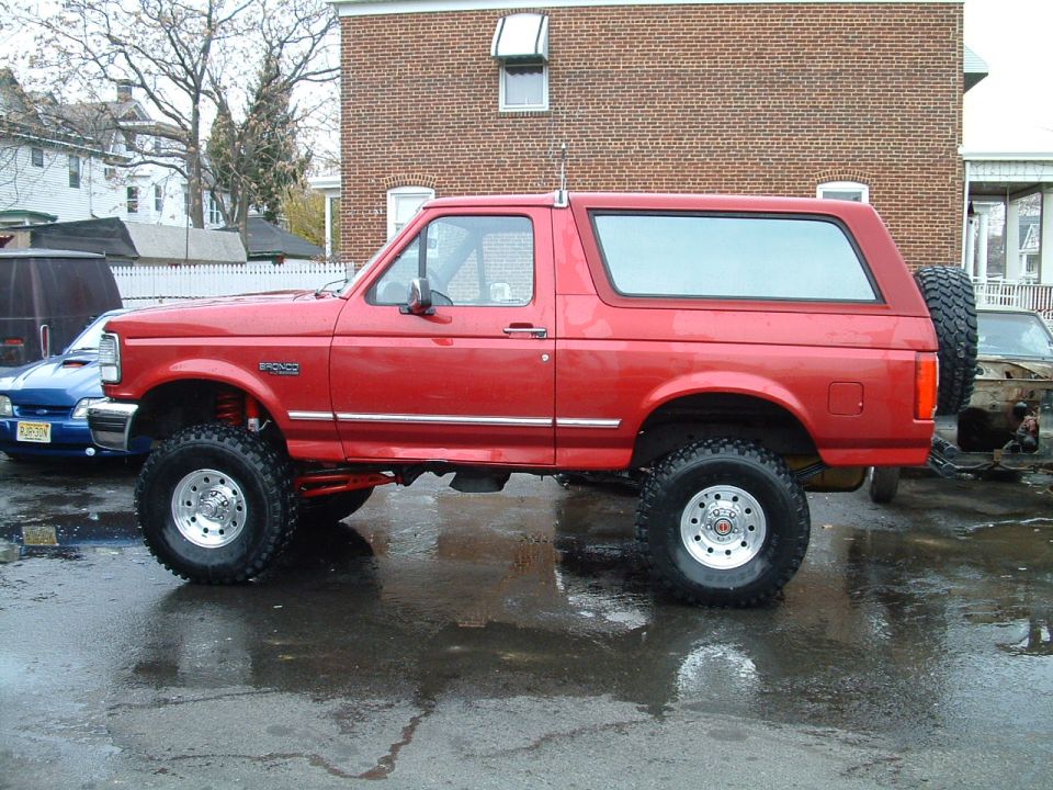 Red lifted 1994 Ford Bronco parked on wet asphalt, another vehicle in background